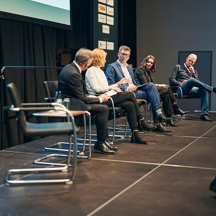 Fünf Personen sitzen auf einer Bühne in einer Podiumsdiskussion. Sie sitzen in einer Reihe auf Stühlen, sprechen miteinander und halten Unterlagen in den Händen. Im Hintergrund sind Sponsorenlogos und eine Leinwand zu sehen.