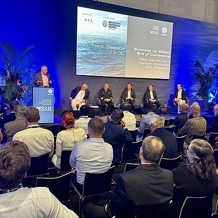 Konferenzraum mit Publikum; auf einer Bühne sitzen mehrere Personen in einer Podiumsrunde, während eine Person am Rednerpult spricht. Im Hintergrund ist eine große Leinwand mit Präsentationsfolien zu sehen.