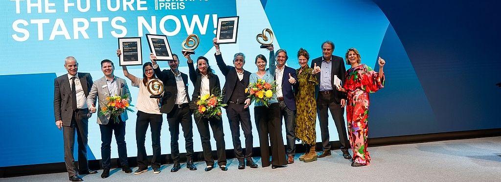 Gewinnerinnen und Gewinner des INTERNORGA Zukunftspreises 2024 jubeln auf der Bühne mit Trophäen, Urkunden und Blumen. Im Hintergrund die Leinwand mit „The Future Starts Now!“