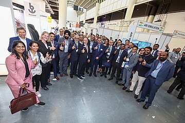 Eine große Gruppe von Menschen steht in einer Messehalle zusammen und posiert für ein gemeinsames Foto. Viele von ihnen zeigen den Daumen nach oben. Im Hintergrund sind Messestände, Firmenbeschilderungen und ein hoher, lichtdurchfluteter Hallenbereich zu sehen. Die Personen tragen überwiegend Businesskleidung und wirken wie Teilnehmende einer Fachmesse oder eines Branchenevents.