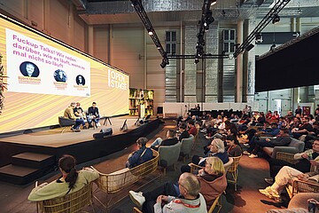 Eine große offene Veranstaltungsfläche mit einer Bühne, auf der zwei Personen in Sesseln sitzen und ein Gespräch vor Publikum führen. Auf einer großen Leinwand hinter ihnen ist der Titel einer Talkrunde zu sehen. Vor der Bühne sitzen zahlreiche Besucherinnen und Besucher in Sesseln und auf Stühlen und hören der Diskussion zu. Die Umgebung wirkt modern, mit Bühnenlicht, Pflanzenwänden und hoher Hallendecke.