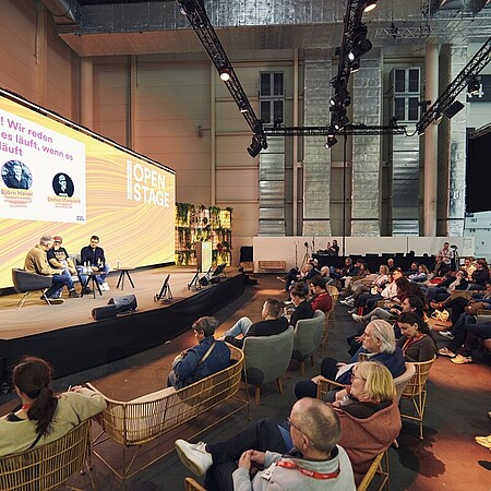 Eine große offene Veranstaltungsfläche mit einer Bühne, auf der zwei Personen in Sesseln sitzen und ein Gespräch vor Publikum führen. Auf einer großen Leinwand hinter ihnen ist der Titel einer Talkrunde zu sehen. Vor der Bühne sitzen zahlreiche Besucherinnen und Besucher in Sesseln und auf Stühlen und hören der Diskussion zu. Die Umgebung wirkt modern, mit Bühnenlicht, Pflanzenwänden und hoher Hallendecke.