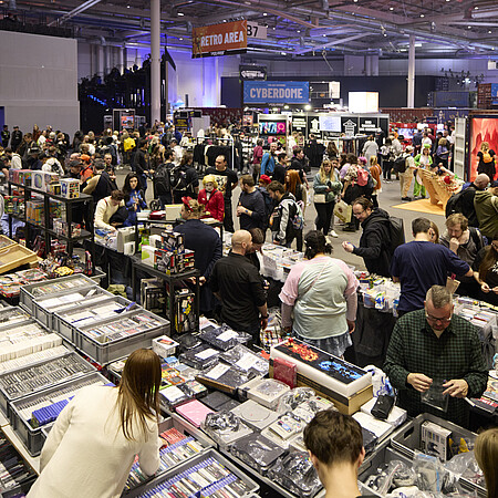 Große Messehalle mit vielen Menschen, die an dicht gefüllten Verkaufsständen vorbeigehen. Auf den Tischen liegen zahlreiche Spiele, Sammelkarten, Figuren und Merchandise-Artikel. Im Hintergrund sind weitere Bereiche der Veranstaltung mit Schildern wie ‚Retro Area‘, ‚Cybedrome‘ und ‚Indie Area‘ zu sehen. Die Halle ist hell ausgeleuchtet und stark besucht.
