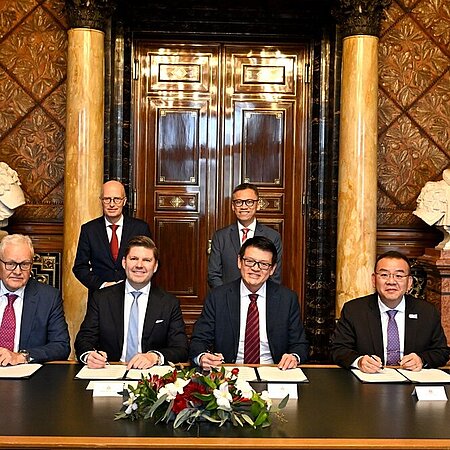 Fünf Personen sitzen an einem Tisch und unterzeichnen Dokumente, dahinter zwei stehende Personen, prunkvoller Raum mit Marmorsäulen und Büsten.