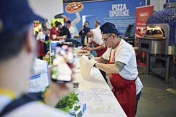 In einer lebhaften Messeszene bereitet eine Person an einem langen Arbeitstisch einen Pizzateig vor. Die Person trägt eine weiße Kleidungsschicht, eine rote Schürze und eine dunkle Kopfbedeckung. Auf dem Tisch liegen Zutaten, Küchenwerkzeuge und ein rundes Gitter zum Abkühlen. Im Vordergrund hält eine andere Person ein Smartphone und filmt oder fotografiert die Szene. Im Hintergrund arbeiten weitere Personen an ähnlichen Stationen. Rechts steht ein großer, blau gekachelter Pizzaofen. Die Umgebung ist farbenfroh gestaltet, mit einem blauen Wandbereich, auf dem „PIZZA EXPERIENCE“ zu lesen ist.