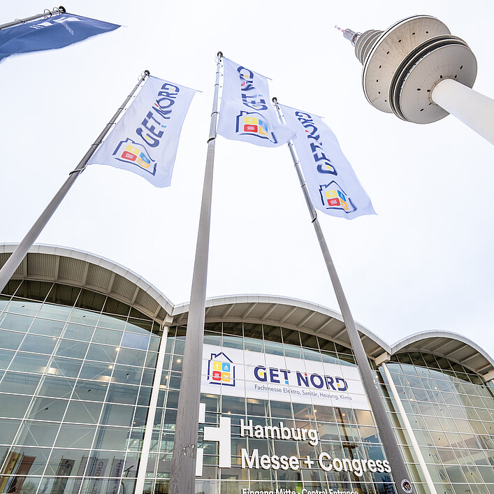 Blick von unten auf mehrere Fahnen mit der Aufschrift „GET NORD“, die vor dem Gebäude der Hamburg Messe + Congress im Wind wehen; im Hintergrund sind die Glasfassade, der Eingangsbereich mit Beschriftung sowie ein hoher Fernsehturm und eine Straßenleuchte zu sehen.