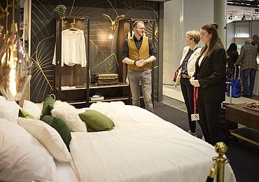 Eine stilvolle Schlafzimmerausstellung mit einem gemütlich dekorierten Bett in Weiß und Grüntönen im Vordergrund. Im Hintergrund steht ein Mann in einer gelben Weste und unterhält sich mit zwei Frauen, die das Design begutachten.