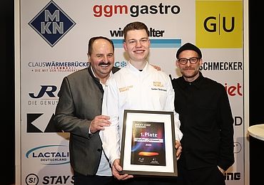 Der Gewinner des Next Chef Awards 2025, Gedion Beckmann, hält stolz seine Siegerurkunde, während Sternekoch Johann Lafer (links) und Moderator Sebastian E. Merget (rechts) ihn gratulierend flankieren.