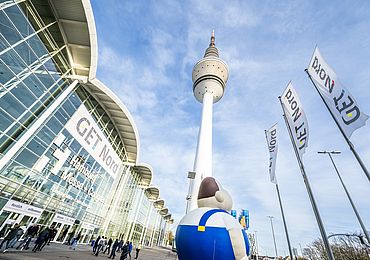 Das Messegebäude ist zu sehen, der Hamburger Fernsehturm und drei Get Nord Flaggen. Personen laufen draußen umher und es ist eine große aufgeblasene Figur zu sehen. 