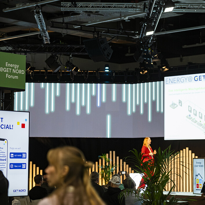 Besuchte Fachveranstaltung auf einer Bühne des Energy@GET NORD Forums. Im Vordergrund sitzen und stehen Teilnehmende, während auf der Bühne eine Präsentation mit großer Leinwand gezeigt wird.