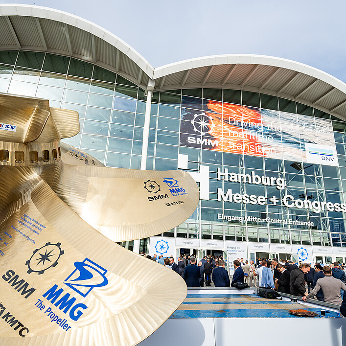 Großes Schiffspropeller-Modell vor dem Eingang der Hamburg Messe. Im Hintergrund die verglaste Fassade des Messegebäudes mit SMM-Banner und zahlreichen Besucherinnen und Besuchern vor dem Haupteingang.