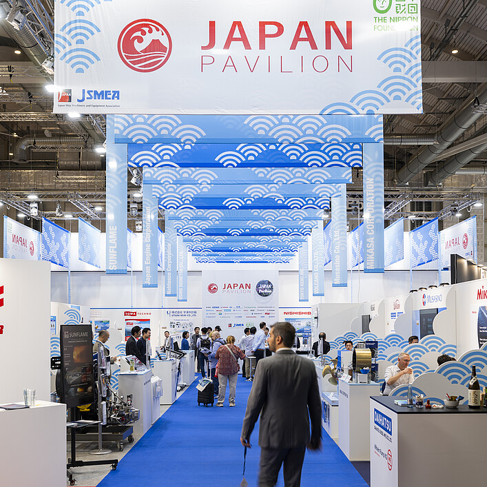 Blick durch den Japan Pavilion in einer Messehalle: Ein blauer Teppich führt durch Ausstellungsstände maritimer Unternehmen, darüber hängen Banner mit Wellenmotiven und der Aufschrift „Japan Pavilion“.