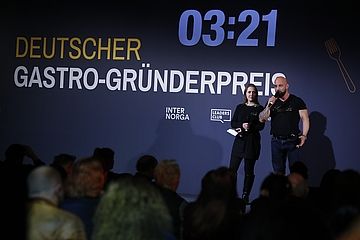 Zwei Menschen stehen auf der Bühne des Deutschen Gastro-Gründerpreises. Im Hintergrund zeigt eine große Leinwand den Eventnamen und einen Countdown.