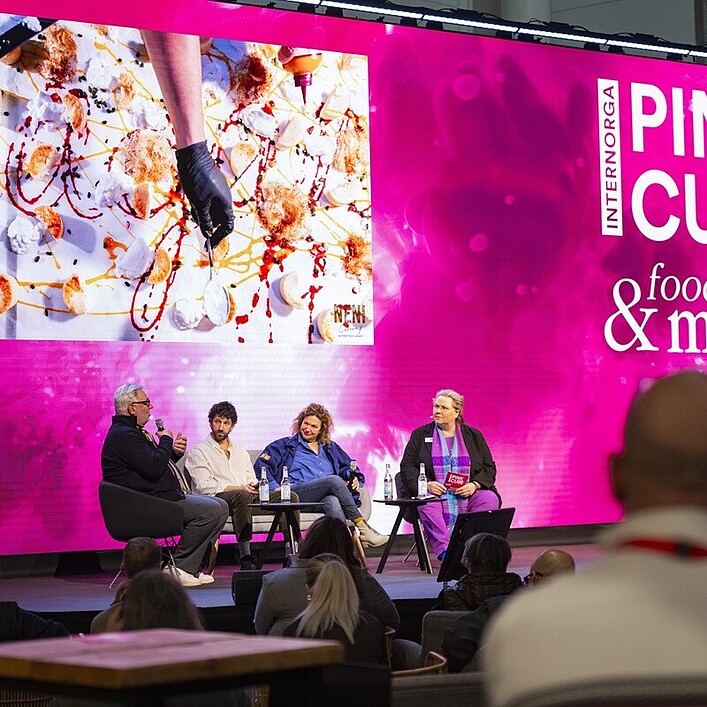 Auf einer Bühne vor einem großen Publikum sitzen vier Personen in Sesseln und führen eine Podiumsdiskussion. Hinter ihnen erstreckt sich eine sehr große, leuchtend pinke LED‑Wand mit dem Schriftzug „INTERNORGA PINK CUBE & food more“. Links auf der Wand ist ein großes Foto eingeblendet, das eine kulinarische Kreation mit verschiedenen Zutaten und farbenfrohen Saucen zeigt. Die Bühne ist mit Mikrofonen und Wassergläsern ausgestattet. Im Vordergrund sind die Silhouetten einiger Zuschauerinnen und Zuschauer zu sehen, die der Diskussion folgen.