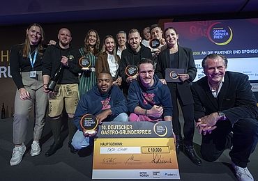 Eine Gruppe von Menschen, darunter die Gewinner von „Taco Craze“, posiert auf der Bühne mit Trophäen und einem großen Scheck über 10.000 Euro für den Deutschen Gastro-Gründerpreis.