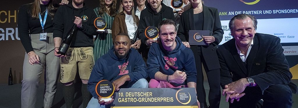 Eine Gruppe von Menschen, darunter die Gewinner von „Taco Craze“, posiert auf der Bühne mit Trophäen und einem großen Scheck über 10.000 Euro für den Deutschen Gastro-Gründerpreis.