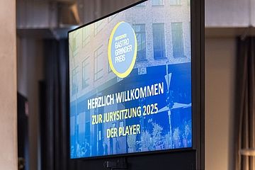 Ein Bildschirm zeigt die Begrüßung zur Jurysitzung 2025 des „Deutschen Gastro Gründerpreises“. Der Text ist in Blau und Gelb gehalten, im Hintergrund des Bildschirms ist eine Stadtszene mit einem Gebäude sichtbar.