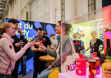 Eine fröhliche Szene auf einer Messe, bei der zwei Frauen mit Gläsern anstoßen. Im Hintergrund sind weitere Personen an einem Stand mit einer farbenfrohen Kulisse und der Aufschrift „Bist du ready?“ zu sehen.