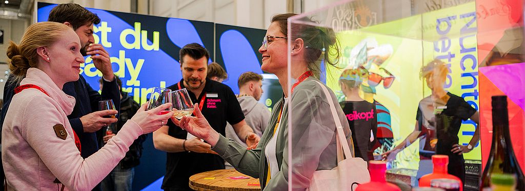 Eine fröhliche Szene auf einer Messe, bei der zwei Frauen mit Gläsern anstoßen. Im Hintergrund sind weitere Personen an einem Stand mit einer farbenfrohen Kulisse und der Aufschrift „Bist du ready?“ zu sehen.