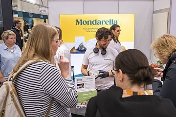 Ein Messestand von „Mondarella“, der pflanzliche Käsealternativen präsentiert. Ein freundlicher Mitarbeiter in einem weißen T-Shirt mit Kopfhörern um den Hals verteilt Kostproben an interessierte Besucher.