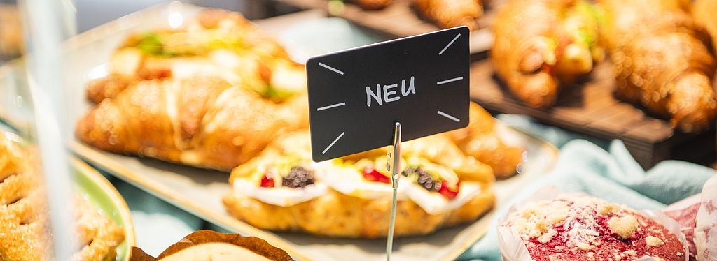 Nahaufnahme einer Auslage in einer Bäckerei mit verschiedenen Backwaren wie Croissants und Muffins. Ein kleines Schild mit der Aufschrift „NEU“ hebt neue Produkte hervor.