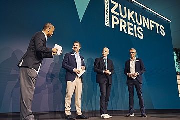 Vier Männer stehen auf einer Bühne vor einer blauen Leinwand mit der Aufschrift "Zukunftspreis" und sind in eine lebhafte Unterhaltung vertieft, während einer von ihnen ein Mikrofon und Karten hält.