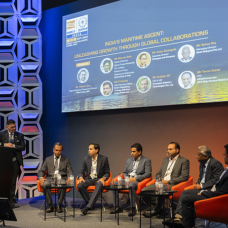 Podiumsdiskussion auf einer Konferenzbühne. Eine Person steht an einem Rednerpult mit der Aufschrift „Open Stage“, während sechs weitere Personen auf orangefarbenen Sesseln sitzen. Im Hintergrund zeigt eine große Leinwand Informationen zu einer maritimen Fachveranstaltung in Indien. Die Bühne ist in Blau- und Orangetönen beleuchtet und von dekorativen Wandpaneelen eingerahmt.