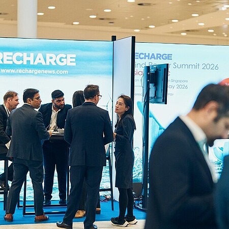 Auf einer Messe stehen mehrere Personen an einem Ausstellungsstand mit dem Schriftzug „RECHARGE“. Der Stand zeigt ein großformatiges Bild von Windkraftanlagen auf dem Meer. Die Personen tragen formelle Kleidung und unterhalten sich in kleinen Gruppen an Stehtischen. Im Vordergrund ist ein weiterer Messestand zu sehen, dessen blaues Display teilweise den Text „Global… for w…“ zeigt. Die Halle ist hell ausgeleuchtet, mit glänzendem Boden und weiteren Ständen im Hintergrund.