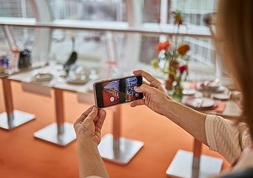 Eine Person fotografiert mit einem Smartphone den „Skywalk“, eine modern gestaltete Fläche mit elegant gedeckten Tischen, floralen Dekorationen und großen Fenstern, die für eine lichtdurchflutete Atmosphäre sorgen.