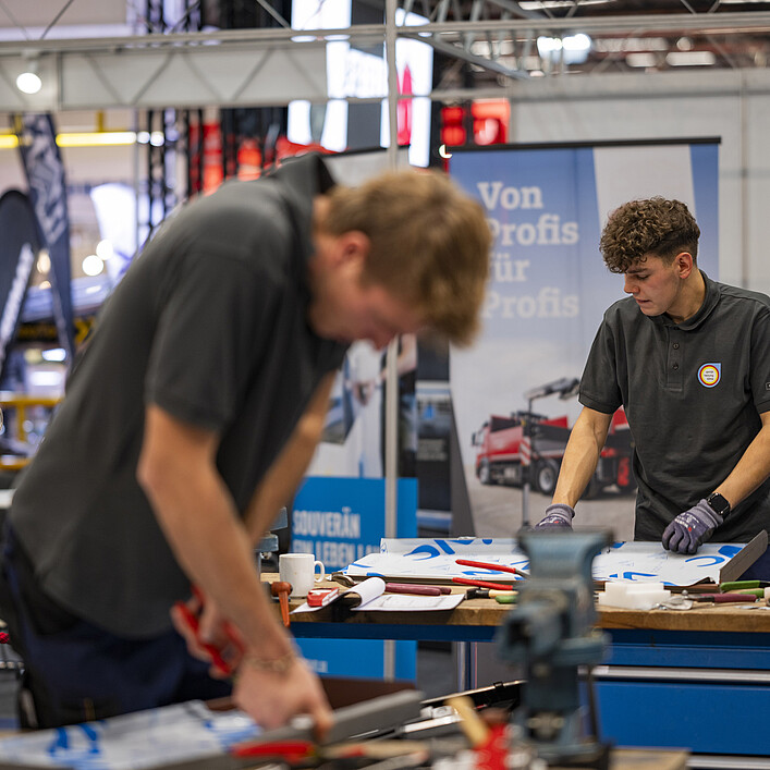 Zwei Personen arbeiten an einer Werkbank in einer Messe- oder Ausstellungshalle. Auf dem Tisch liegen Werkzeuge und bearbeitete Blechteile. Im Vordergrund ist eine Person unscharf bei der Metallbearbeitung zu sehen, während eine weitere Person an einem Werkstück arbeitet. Im Hintergrund befinden sich Maschinen, Messestände und ein Banner mit der Aufschrift „Von Profis für Profis“.