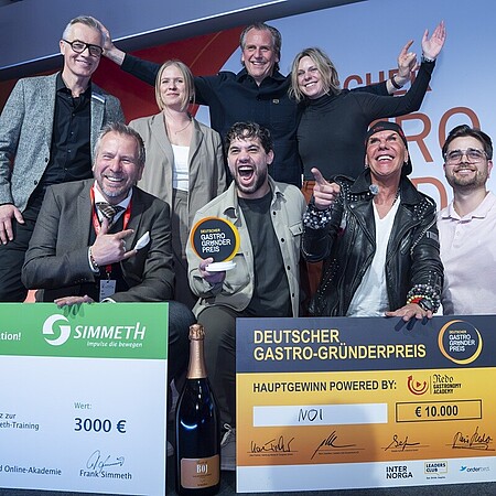 Eine Gruppe von neun Personen posiert auf einer Bühne vor einem großen orange‑weißen Hintergrund mit der Aufschrift „Deutscher Gastro‑Gründerpreis“. Vor der Gruppe stehen zwei große symbolische Schecks: links ein grüner Scheck über 3.000 Euro für einen Ausbildungsplatz bei Simmet‑Training, rechts ein orangefarbener Hauptgewinn‑Scheck über 10.000 Euro mit dem Gewinnernamen „MOI“. Davor steht zudem eine große Flasche Sekt. Eine Person in der Mitte hält eine runde Auszeichnungstrophäe hoch. Die Gruppe zeigt teils jubelnde Gesten und wirkt wie bei einer Siegerehrung. Geben Sie Ihr Feedback zu BizChat ab.
